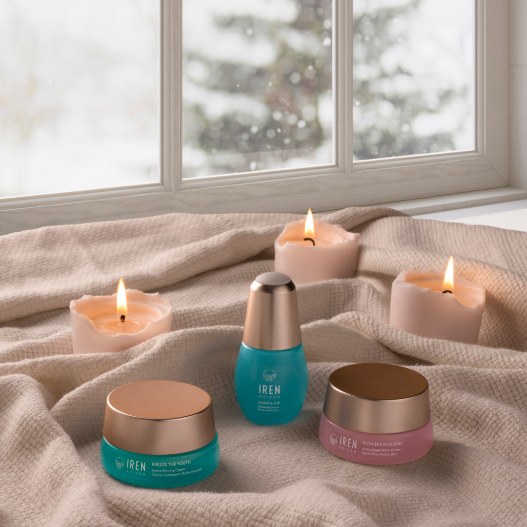 The MOCHI SKIN Instant Glow Travel Set by Fast Bundle and three lit candles are arranged on beige fabric in front of a window with a snowy outdoor view.