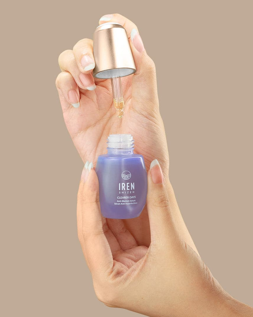 A hand holds a blue glass dropper bottle labeled "IREN Shizen," identifying it as the CLEARER DAYS Anti-Blemish Serum. The dropper, lifted from the bottle, drips clear liquid—a potent solution—against a plain beige background.