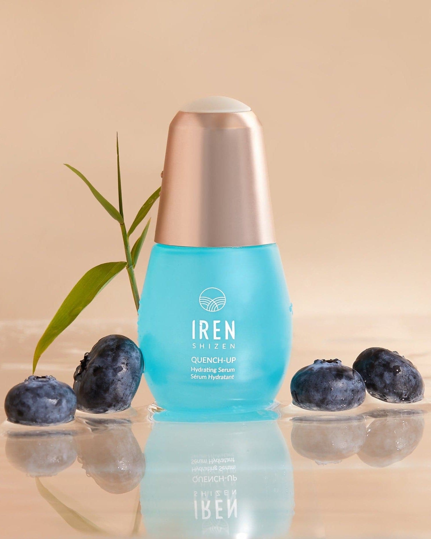 A blue bottle of QUENCH-UP Hydrating Serum by IREN Shizen stands on a reflective surface, enriched with Hyaluronic Acid and surrounded by blueberries and a green leaf.