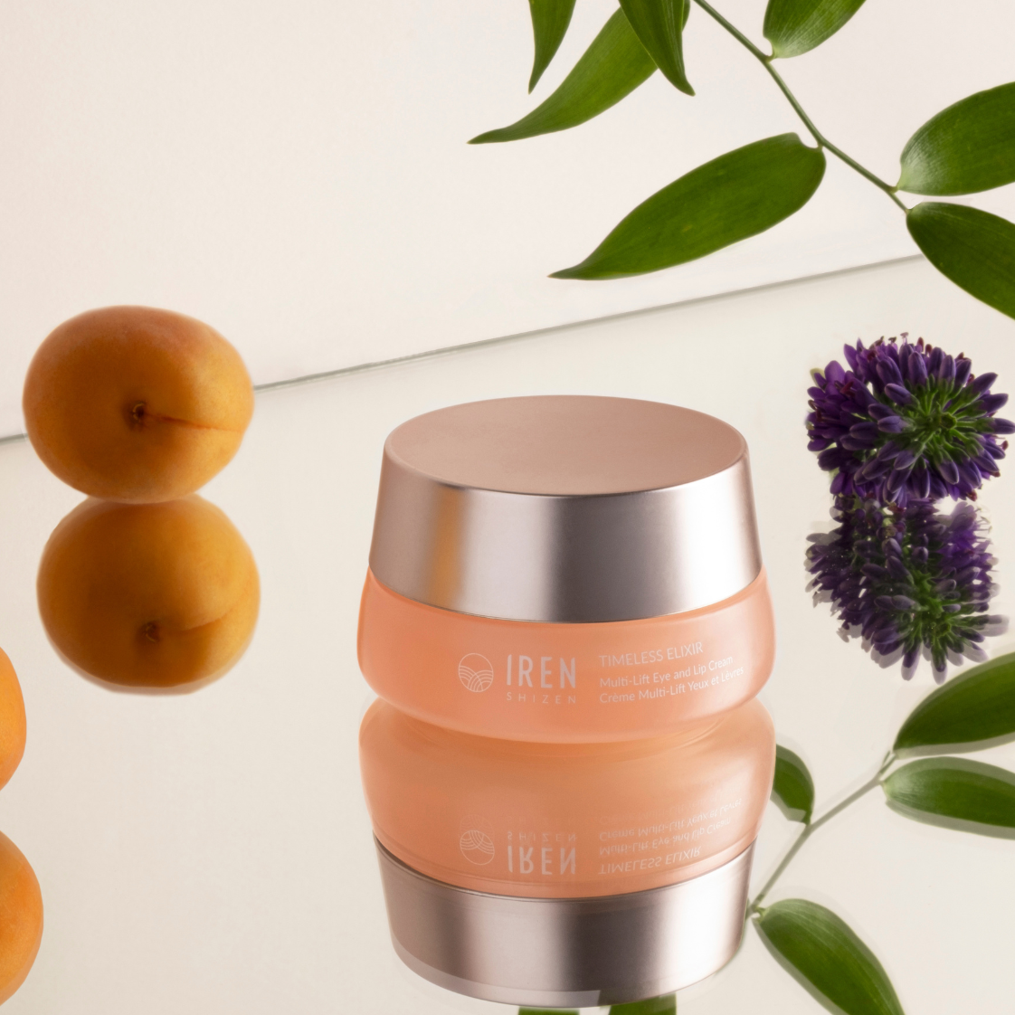 A jar of IREN Shizen TIMELESS ELIXIR Hydrating Eye Skincare Kit, celebrated for its anti-aging benefits, is placed on a reflective surface, surrounded by apricots and green leaves, with a purple flower nearby.