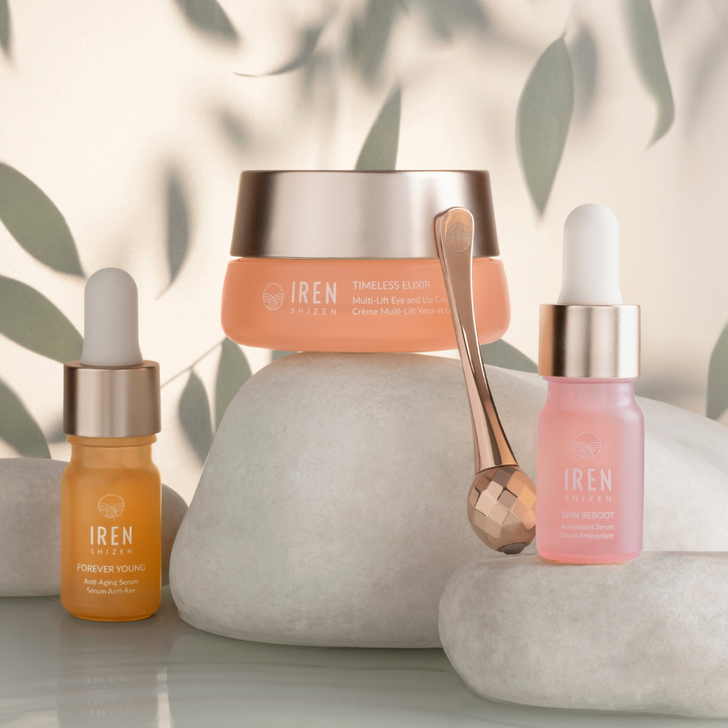 Skincare products including a jar, two dropper bottles, and a small facial roller—all with gold accents—are elegantly displayed on white stones with leaves casting shadows in the background. Among them is the TIMELESS ELIXIR Anti-Aging Eye Skincare Kit by IREN Shizen, an anti-aging eye treatment designed for dark circles reduction.