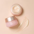 A jar of pink-colored face cream with a gold lid is open, revealing the cream inside. Next to it lies a smear of the cream on a beige surface. The jar, which is part of the special PINK OCTOBER Skincare Set, is labeled "IREN Shizen" and "FLOWERS BELOVED.