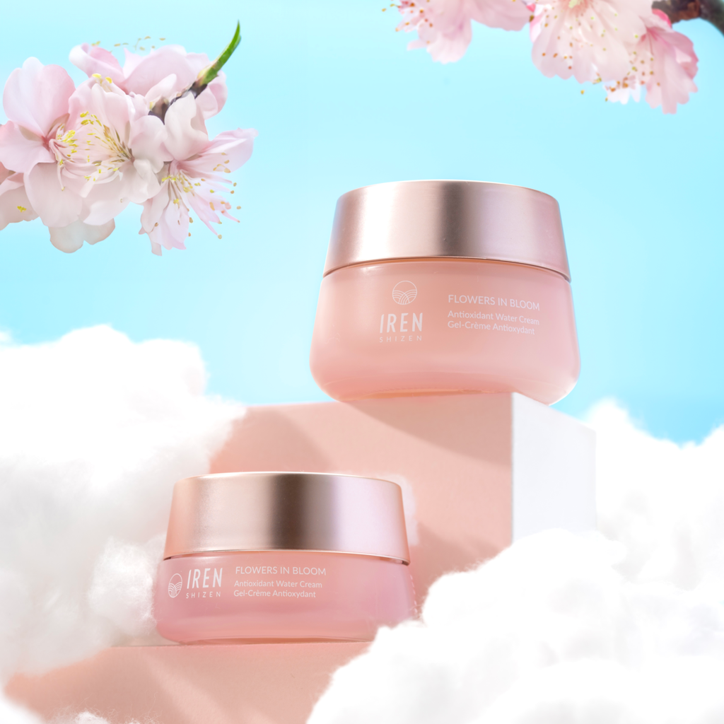 Two jars of IREN Shizen's FLOWERS IN BLOOM Antioxidant Water Cream, a silicone-free gel cream, are displayed on clouds with cherry blossoms against a clear blue sky background.