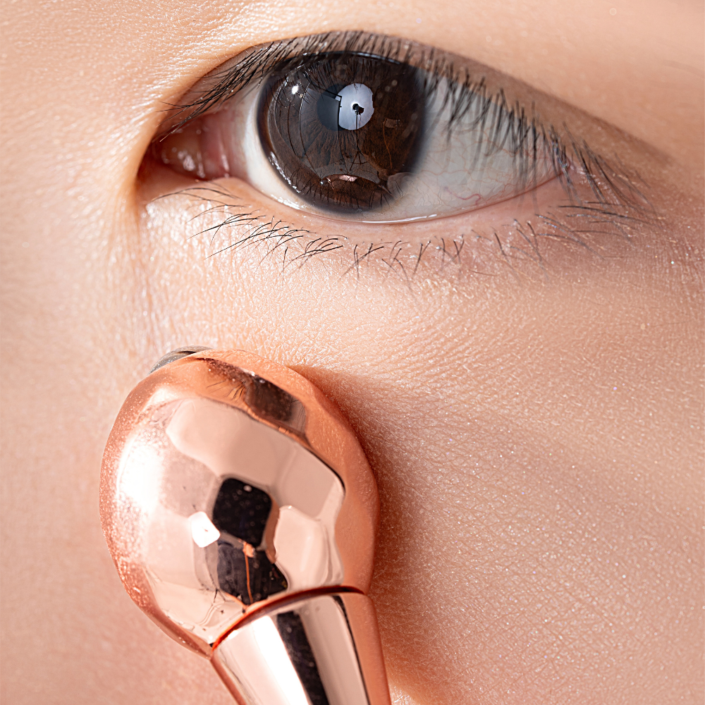 Close-up of a person's eye with the rose gold facial massage tool being applied to the skin underneath, targeting dark circles with IREN Shizen's TIMELESS ELIXIR Multi-Lift Eye and Lip Cream.