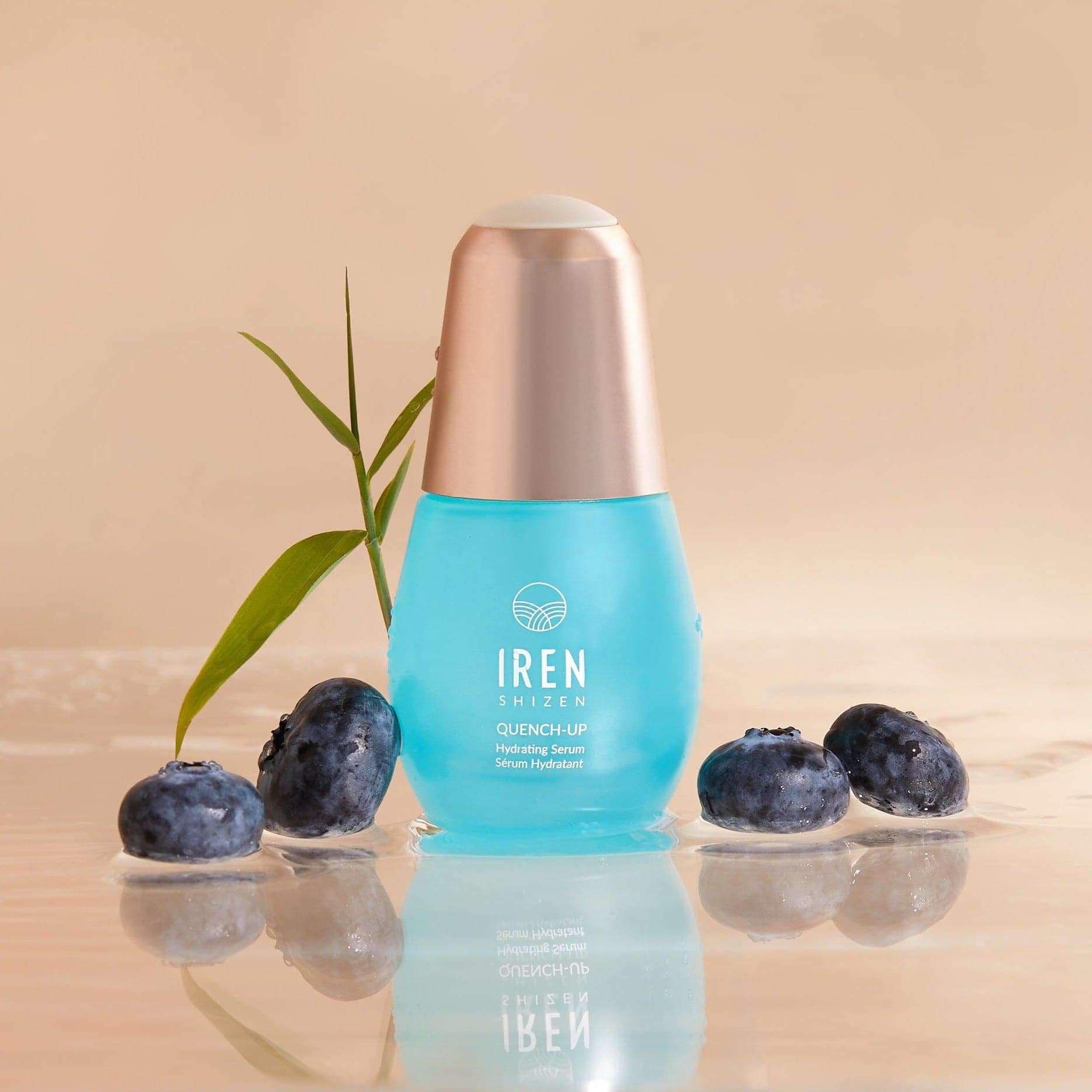 A blue bottle of QUENCH-UP Hydrating Serum by IREN Shizen stands on a reflective surface, enriched with Hyaluronic Acid and surrounded by blueberries and a green leaf.